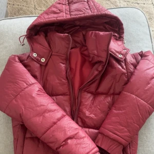 Vinröd pufferjacka med huva - Säljer en vinröd pufferjacka med huva och dragkedja. Jackan är quiltad och har ribbade muddar vid ärmslut och nederkant. Perfekt för kalla dagar och ger en snygg, sportig look. Passformen är normal och jackan är lätt att matcha med jeans eller leggings.