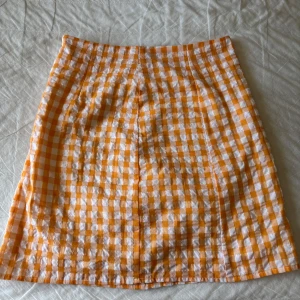 Rutig kortkjol  - Superfin kortkjol i orange och vitt rutigt mönster. Kjolen har en rak passform. Den stängs med en vit dragkedja i sidan och är perfekt för sommarens alla tillfällen.