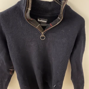 Mörkblå Barbour stickad tröja half zip - Snygg mörkblå stickad tröja från Barbour med half zip och ringformad dragkedja. Tröjan har långärmade ärmar och klassisk krage med rutigt foder. Perfekt för dig som gillar stilren och tidlös design. Ny pris 1800