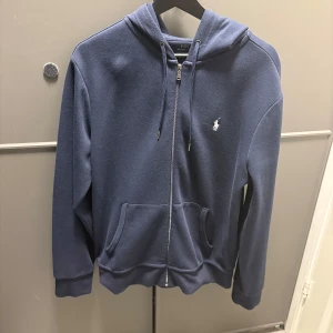  Ralph Lauren zip  hoodie - Snygg marinblå hoodie från Polo Ralph Lauren med klassisk broderad logga på bröstet. Tröjan har huva med snörning, dragkedja framtill och två praktiska fickor. Tillverkad i mjuk bomull som är skön att ha på sig. Perfekt för chill dagar och streetstyle.