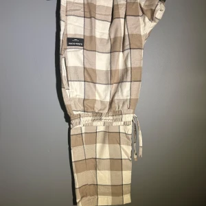 Rutiga byxor från Björn Borg - Beige och bruna rutiga byxor från Björn Borg med elastisk midja och snörning. Lätta och sköna, perfekta för chill dagar. Mjukt bomullsmaterial och avslappnad passform. Snyggt rutmönster i neutrala färger.