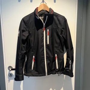 Helly Hansen jacka  - Vindjacka från Helly Hansen med vita dragkedjor och logga på ärmen och ryggen. Den är fodrad så hyfsat varm och tål regn bra. Storlek XS passar s
