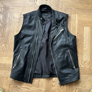 Svart skinnväst från Diesel - Snygg svart skinnväst från Diesel med cool bikerstil. Västen har flera silvriga dragkedjor, ståkrage med tryckknapp och en stilren, rak passform. Perfekt att slänga över en hoodie eller t-shirt för en edgy look.