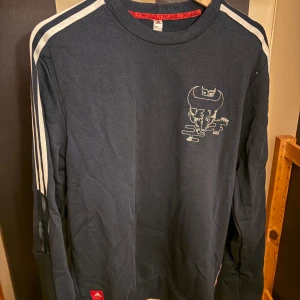 Adidas Arsenal långärmad tröja marinblå - Marinblå långärmad tröja från Adidas x Arsenal med vita ränder längs axlarna, tryckt grafik på bröstet och röd etikett nertill. Skön passform och klassisk sportig look, perfekt för dig som gillar fotboll och streetwear.