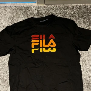 Svart Fila t-shirt med färgglad logga - Svart t-shirt från Fila med stor, färgglad logga i rött, orange och gult framtill. Klassisk passform och rund halsringning. Perfekt för dig som gillar streetwear och vill sticka ut med en snygg grafisk detalj.
