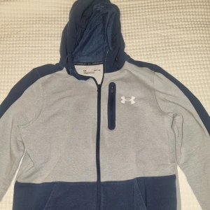 Under Armour hoodie i grått och blått - Hoodie från Under Armour i grått och marinblått med dragkedja framtill och bröstficka med dragkedja. Vit logga på bröstet och huva med dragsko. 