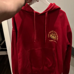 Röd hoodie Vaillent med tryck M - Röd hoodie från Vaillent i storlek M med stor färgglad smiley och texten 'Eat your vitamins, happy inside' på ryggen. Tröjan har huva med snörning, magficka och är tillverkad i mjuk bomull. Passar m, är i bra skick. Enda som är ”fel” är att den tappat lite färg på bottnen av ett av snörena. Köpt för 899 kr