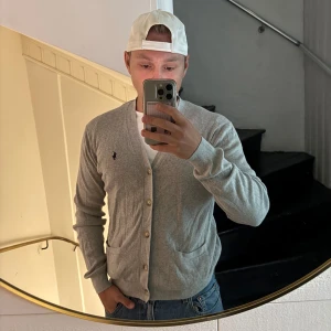 Grå kofta från Polo Ralph Lauren - Snygg ljusgrå kofta från Polo Ralph Lauren med klassisk v-ringning, knappar framtill och två fickor. Ikoniska loggan broderad på bröstet. Perfekt att slänga över en t-shirt för en clean och chill look.