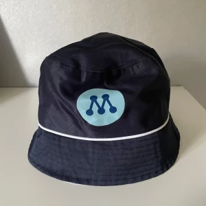 Moderaterna bucket hat - Bucket hat i mörkblått med vit kant och moderaternas logga framtill. Hatten har en klassisk rund form och är tillverkad i ett syntetmaterial som känns lätt och smidigt. Insidan av brättet är vit och har en diskret textur. Perfekt för dig som vill sticka ut med en cool accessoar.