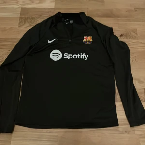 Svart långärmad FC Barcelona Nike-tröja - Svart långärmad fotbollströja från Nike med FC Barcelona-emblem på bröstet och Spotify-logga framtill. Tröjan har halv dragkedja vid halsen och är tillverkad i ett lätt, sportigt material som passar perfekt för träning eller match.