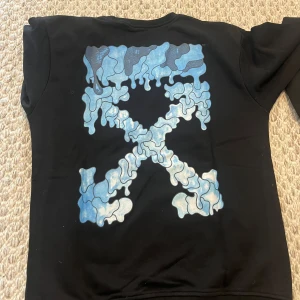 Off-White Sweatshirt - Svart t-shirt från Off-White med korta ärmar och rund hals. På framsidan finns ett vitt Off-White-tryck med handmotiv, och på ryggen ett stort blått grafiskt tryck i form av Off-White-loggan. Materialet är mjuk bomull och passformen är normal.