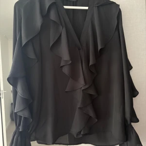 Svart volangblus från Zara XS - Supersnygg svart blus från Zara i storlek XS med stora volangdetaljer längs ärmar och framtill. Blusen har v-ringning och långa ärmar med resår vid ärmslut. Perfekt för dig som gillar en trendig och chic look.