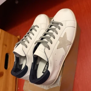 Golden Goose Superstar vita sneakers - Golden Goose Superstar sneakers i vitt skinn med grå mockastjärna på sidan, grå snörning och mörkblå häl. Klassisk låg modell med rund tå och platt sula. Snygga detaljer och ikonisk design – perfekta för dig som gillar streetstyle.