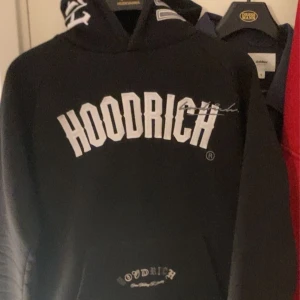 Svart HOODRICH hoodie med tryck - Svart hoodie från Hoodrich med stora vita tryck på bröstet och huvan. Klassisk passform med lång ärm och huva med feta bokstäver. Perfekt streetstyle-plagg med coola detaljer och skön känsla. Materialet är mjukt och passar till lager-på-lager.
