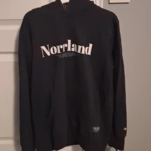 Svart hoodie Norrland SQRTN Company XS - Svart hoodie från SQRTN Company i storlek XS med vit Norrland-tryck på bröstet. Klassisk modell med huva och känguruficka framtill. Perfekt för dig som gillar streetwear och vill visa din norrländska pride.