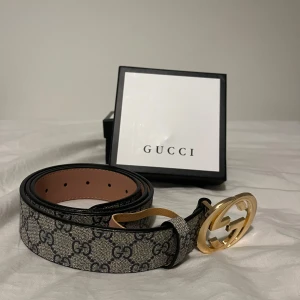 Guccci bälte med guldspänne - Snyggt Guccci -bälte i klassiskt GG Supreme-mönster med beige och svarta toner. Bältet har ett stort, rundat guldspänne med dubbla G-loggan och är tillverkat i exklusivt skinn. Perfekt accessoar för att lyfta din outfit med lyxig känsla.