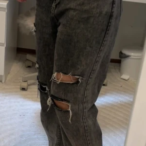 Gråa slitna jeans - Säljer ett par gråa jeans med snygga slitningar över knäna. Jeansen har en cool, tvättad look och passar perfekt till en avslappnad streetstyle. Materialet är klassisk denim och modellen är loose fit.