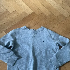 Grå sweatshirt från Polo Ralph Lauren - Klassisk grå sweatshirt från Polo Ralph Lauren med rund halsringning och broderad marinblå logga på bröstet. Tröjan har långa ärmar, ribbade muddar och är tillverkad i mjuk bomullsmix. Perfekt för en chill och stilren look.