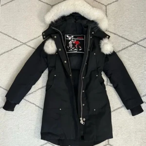 Svart dunjacka Moose Knuckles XS - Snygg svart dunjacka från Moose Knuckles i storlek XS. Jackan har lång passform, avtagbar huva med vit fuskpäls och coola detaljer som dragkedja och knappar. Perfekt för kalla vinterdagar och har en broderad hockeypatch på insidan.