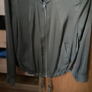 Olivgrön bomberjacka i storlek 38 - Snygg olivgrön bomberjacka med dragkedja framtill och ribbad krage. Jackan har två fickor på sidorna och dragsko nertill för en schysst passform. Perfekt för dig som gillar en clean och stilren look.