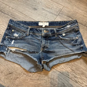 Blå jeansshorts från MNG, strl 36 - Snygga blå jeansshorts från MNG med slitna detaljer och råa kanter. Klassisk femficksmodell med knapp och dragkedja framtill. Shortsen har låg midja och är gjorda i robust denim med coola slitningar för en avslappnad vibe.