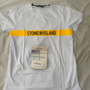 Stone Island vit t-shirt med gul logga - Vit t-shirt från Stone Island med en bred gul rand över bröstet och svart logotyptryck. Klassisk rund hals och korta ärmar. Materialet är mjuk bomull som känns skön mot huden. Perfekt för dig som gillar streetwear och vill sticka ut med en ikonisk design.
