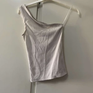 Ljusbeige one shoulder topp - Snygg ljusbeige topp med one shoulder-design och smal axelrem på ena sidan. Ribbad struktur ger en trendig känsla och toppen är figurnära. Perfekt för dig som gillar minimalistisk stil och vill sticka ut lite extra.