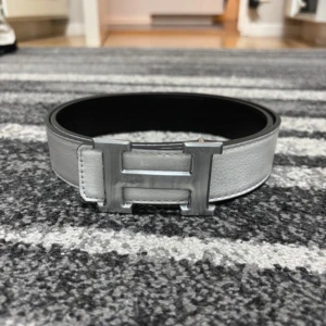  vändbart bälte i skinn - Snyggt vändbart -bälte i skinn med klassiskt H-spänne i silver. Ena sidan är vit och den andra svart, vilket gör det enkelt att matcha med olika outfits. Bältet har en stilren och exklusiv känsla med grov struktur på lädret och bred design.
