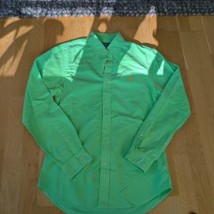 Grön skjorta från Polo Ralph Lauren - Fräsch mintgrön skjorta från Polo Ralph Lauren i slim fit med klassisk button-down krage och orange broderad logga på bröstet. Skjortan är långärmad, har vita knappar och är tillverkad i mjuk bomull. Perfekt för dig som gillar stilrena och färgglada plagg. Axelbredd 48 cm, längdmått 75 cm