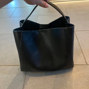 Svart handväska från Gina Tricot - Stilren svart handväska från Gina Tricot i slätt skinnimitation. Väskan har en boxig form, ett stadigt handtag och ett stort huvudfack med innerficka som stängs med dragkedja. Tillhörande längre band som fästes vid sidorna på väskan 