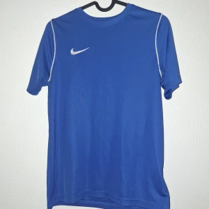 Blå Nike Dri-Fit fotbollströja  - Snygg blå fotbollströja från Nike i Dri-Fit material som andas och håller dig torr under träningen. Tröjan har korta ärmar, vit Nike-logga på bröstet och vita sömmar längs axlarna. Perfekt för dig som vill ha en stilren och sportig look på planen.