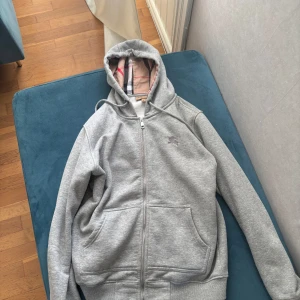 Helt ny Grå hoodie från Burberry med dragkedja - Säljer en grå hoodie från Burberry i storlek M med klassiskt rutigt foder i huvan och insida. Tröjan har broderad Burberry-logga på bröstet, snörning i huvan och två stora fickor framtill. Materialet är mjukt och fleecefodrat för extra komfort.