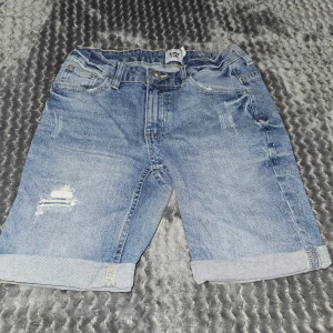 Blå jeansshorts från 157 - Snygga blå jeansshorts från 157 med klassisk femficksdesign, uppvikta benslut och slitna detaljer framtill. Shortsen har normal passform och är tillverkade i mjukt denimtyg med knapp och dragkedja i midjan. Perfekta för en chill sommardag.