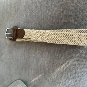 Beige flätat bälte med bruna detaljer - Snyggt beige flätat bälte med bruna skinn-detaljer vid spännet och en klassisk silverfärgad metallspänne. Bältet har en mjuk och flexibel känsla tack vare det vävda materialet och passar perfekt till jeans eller chinos.