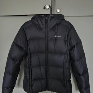 Svart dunjacka Sail Racing M - Svart pufferjacka från Sail Racing i storlek M. Jackan har huva, dragkedja framtill och är fylld med RDS-certifierat ankdun. Yttermaterialet är nylon och jackan är quiltad för extra värme. Perfekt för kalla dagar när du vill hålla stilen.