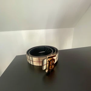 Rutig Burberry bälte med guldspänne - Snyggt bälte från Burberry med klassiskt rutigt mönster i beige, svart, grått och rött. Bältet har ett stort guldigt 'B'-spänne framtill och är gjort i skinn. Perfekt accessoar för att lyfta din outfit med en lyxig touch.