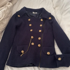 Mörkblå stickad skepparkavaj med guldknappar - Snygg mörkblå stickad skepparkavaj med dubbla rader guldknappar och cool kedjedetalj. Jackan har bred krage, fickor med lock och dekorativa knappar även vid ärmslut. Perfekt för dig som vill ha en unik och trendig look.