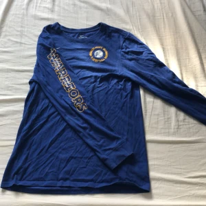 Blå långärmad Golden State Warriors Nike tröja - Golden State Warriors långärmad baskettröja från Nike i klar blå färg. Tryck med Warriors-logga på bröstet och stor text på ena ärmen. Rund hals och mjukt, lätt material som andas. Perfekt för dig som gillar basket och sportig stil. Storleken är Childrens L men vuxen S