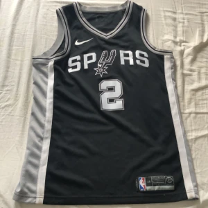 San Antonio Spurs Leonard #2 baskettröja - Autentisk, Snygg svart baskettröja från Nike med San Antonio Spurs tryck och nummer 2, Leonard på ryggen. Tröjan är ärmlös med vita och grå detaljer längs sidorna och kragen. Materialet är lätt och ventilerande polyester, perfekt för sport eller streetwear.