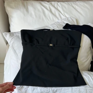 Svart axelbandslös topp från Zara - Säljer en svart axelbandslös topp från Zara med tre smala band och silvriga spännen framtill. Toppen har en snygg, figurnära passform och öppen rygg med banddetaljer. Perfekt att styla med jeans eller kjol för en cool look.