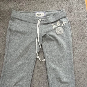 Abercrombie and fitch byxor - Grå mjukisbyxor från Abercrombie & Fitch med broderad A&F-logga och emblem på höften. Byxorna har dragsko i midjan och är tillverkade i mjukt bomullsmaterial. Perfekta för chill dagar och har en relaxed passform.
