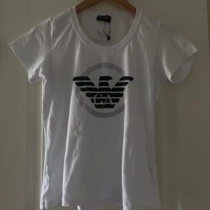 Vit Armani t-shirt med logga - Vit t-shirt från Armani med stor svart logga framtill. Klassisk rund hals och korta ärmar. Materialet är mjuk bomull och den är i mycket bra skick. Storlek XL men passformen är som XS / S