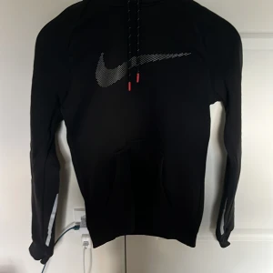 Svart Nike hoodie med reflexdetaljer XS - Svart hoodie från Nike i storlek XS med stor vit logga på bröstet och reflexdetaljer på ärmarna. Tröjan har huva med snörning och en klassisk magficka. Perfekt för dig som gillar sportig och clean stil.
