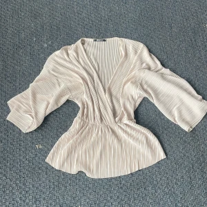 plisserad omlottblus från Gina Tricot - Säljer en superfin ljusbeige omlottblus från Gina Tricot i storlek XS. Blusen har tunna vertikala ränder, v-ringning och vida trekvartsärmar. Materialet är glansigt och känns lyxigt, perfekt för att lyfta din outfit. Tryck ej köp nu