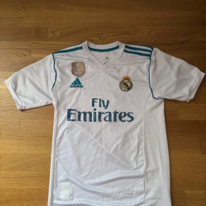 Real Madrid Ronaldo 7 Adidas tröja - Säljer en vit Real Madrid matchtröja från Adidas med Ronaldo 7 på ryggen. Tröjan har korta ärmar, blå detaljer och klubbmärke på bröstet. Materialet är lätt och andas, perfekt för fotboll eller att bära som supporter. Fly Emirates-logga framtill och La Liga-märke på ärmen.