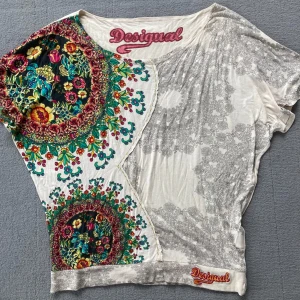 Desigual blommig topp med tryck - Unik topp från Desigual med asymmetrisk design, färgglada blommönster och gråvita detaljer. Stor Desigual-logga fram och broderad nertill. Tunn, mjuk bomullskänsla och kort ärm. Perfekt för dig som gillar färg och mönster! Storleken är som en S/M.