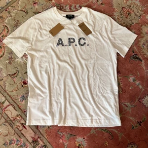 Vit t-shirt från A.P.C. med print - Vit t-shirt från A.P.C. med svart paisleyinspirerat A.P.C.-tryck på bröstet. Klassisk rund hals, korta ärmar och mjuk bomullskvalitet. En clean och stilren tee som lyfter din outfit med en cool detalj framtill.