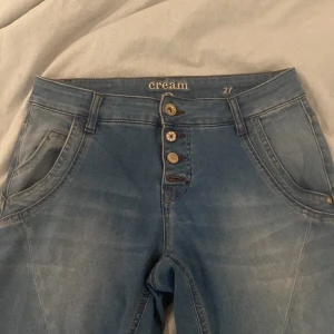 Blå jeans från Cream - Snygga blå jeans från Cream med medium midja och dekorativa knappar framtill. Jeansen har klassisk femficksdesign och är tillverkade i stretchigt denim för en bekväm passform. Perfekta till sneakers eller boots.