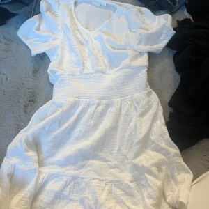 Vit sommarklänning från Gina Tricot - Superfin vit klänning från Gina Tricot med v-ringning, korta ärmar och smock i midjan. Klänningen har en luftig och flowy känsla, perfekt för varma dagar. Tillverkad i mjuk bomull som känns skön mot huden.