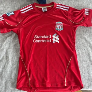 Liverpool FC röd matchtröja Adidas - Liverpool FCs officiella matchtröja i rött med vita Adidas-ränder på axlarna och guldiga detaljer. Broderat klubbmärke på bröstet och Standard Chartered-tryck på framsidan. Tillverkad i lätt och ventilerande polyester, perfekt för fotboll eller supporterliv.